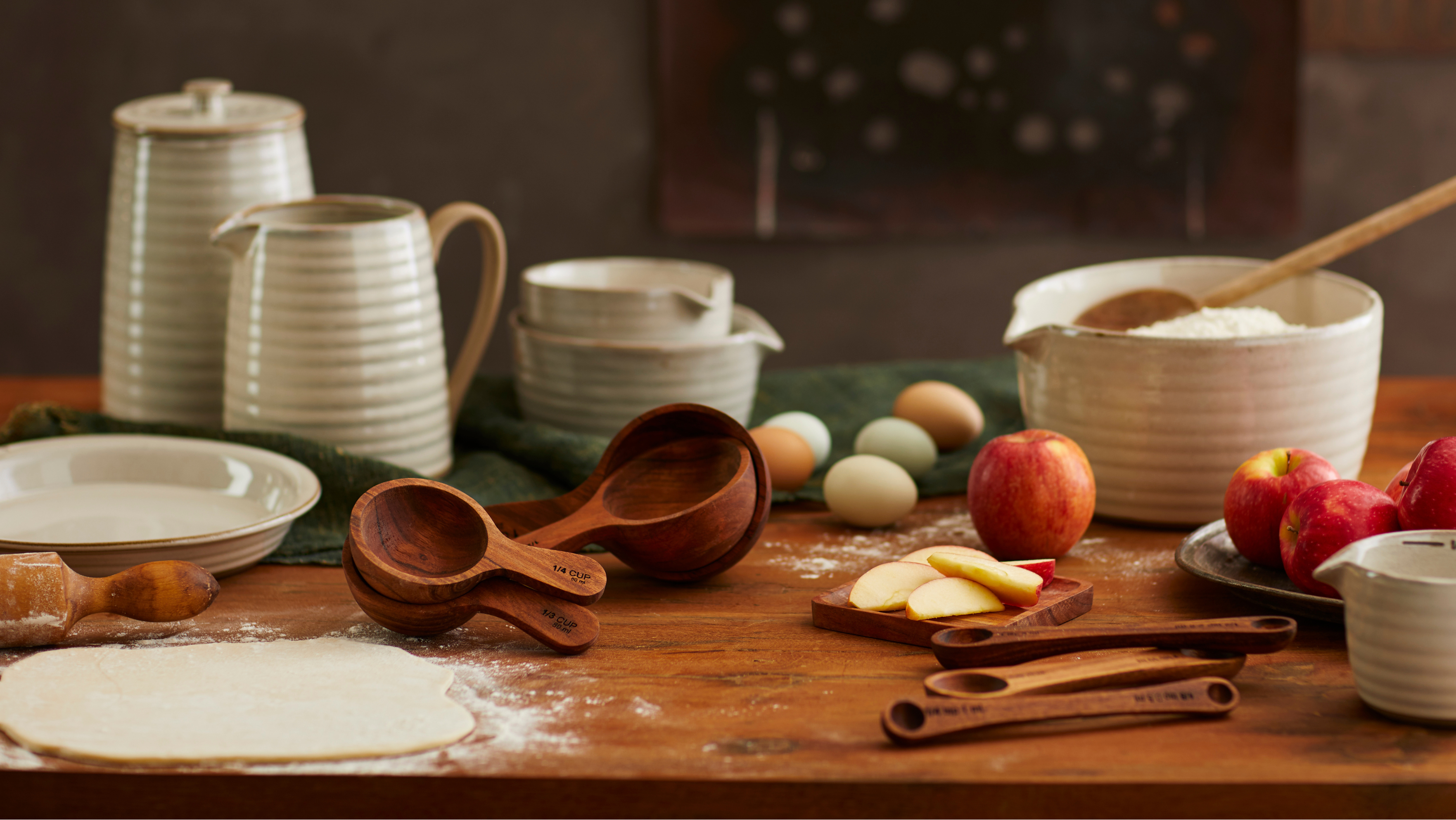 A rustic kitchen scene featuring a coordinated set of cream-colored ribbed ceramic pitchers, bowls, and cups arranged on a wooden table alongside baking ingredients like flour, eggs, apples, and wooden measuring tools.