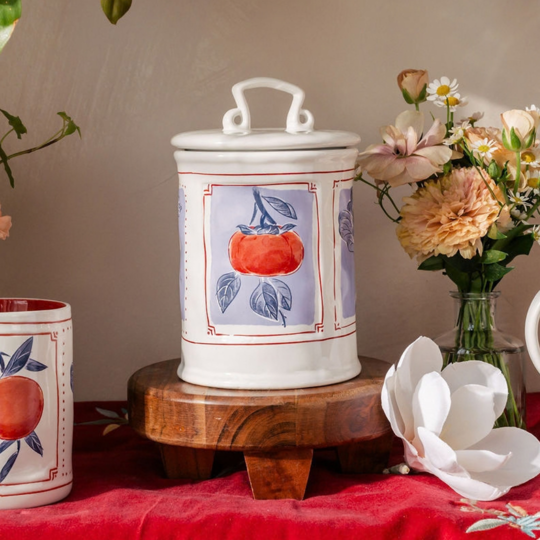 A white ceramic canister with a lid and handle, decorated with a painted persimmon illustration on a lavender panel, displayed on a wooden stand with flowers and a red cloth in the background.