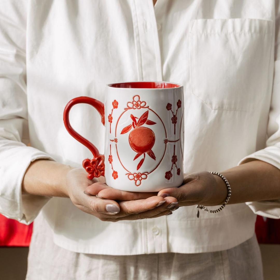 A person in a white button-up shirt holds a white ceramic mug with a red handle, red interior, and a red fruit design in the center.
