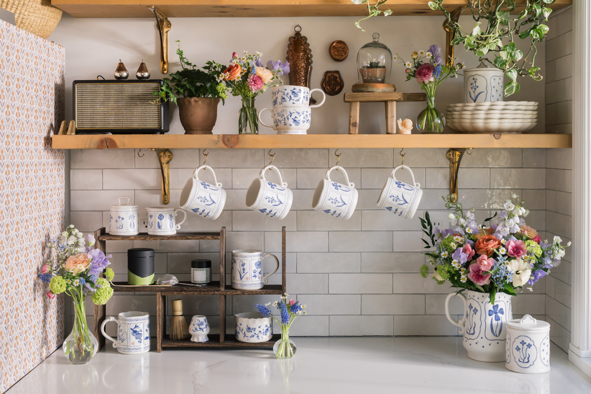 A styled kitchen shelf display featuring multiple white ceramic mugs and pitchers with blue floral patterns, hanging from brass hooks and arranged on wooden shelves alongside flowers, plants, and decorative objects.