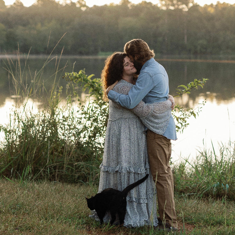 A couple embracing outdoors by a lake at sunset, with a black cat walking nearby on the grass. The woman wears a light blue-gray tiered maxi dress and the man wears a light blue denim shirt and tan pants.
