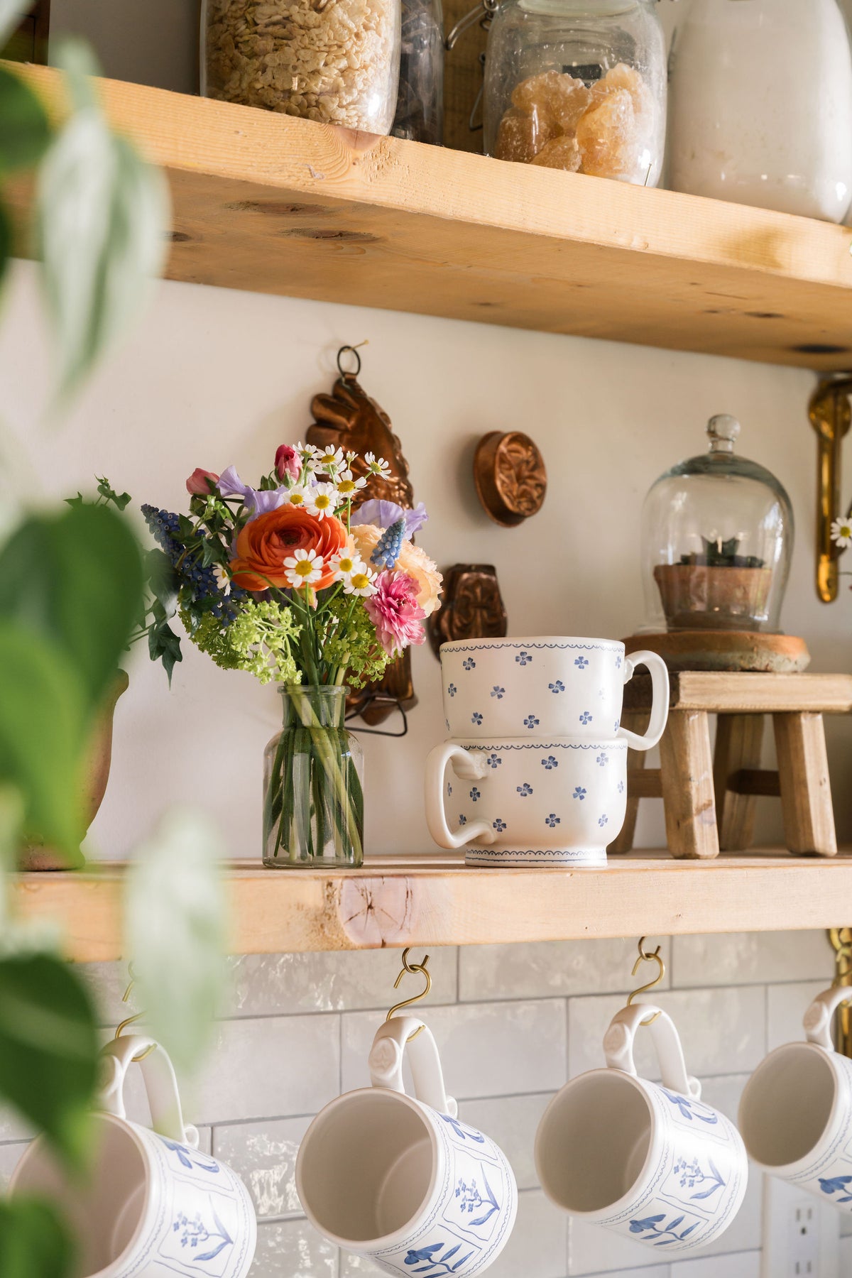 A styled kitchen shelf scene with wooden floating shelves, white ceramic mugs with blue patterns, a glass vase of colorful flowers, and jars on a shelf, photographed in a real interior setting.