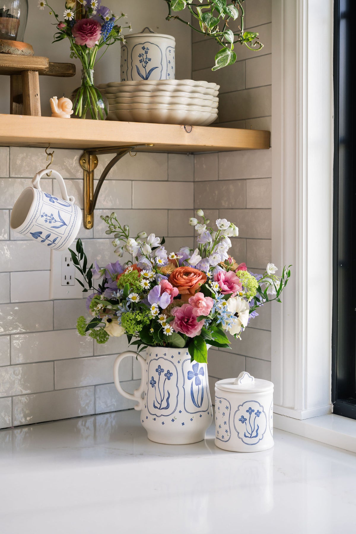 A styled kitchen scene with a white ceramic pitcher and matching lidded jar decorated with blue floral motifs, filled with a colorful bouquet of flowers, set against a white subway tile backsplash and wooden shelf.