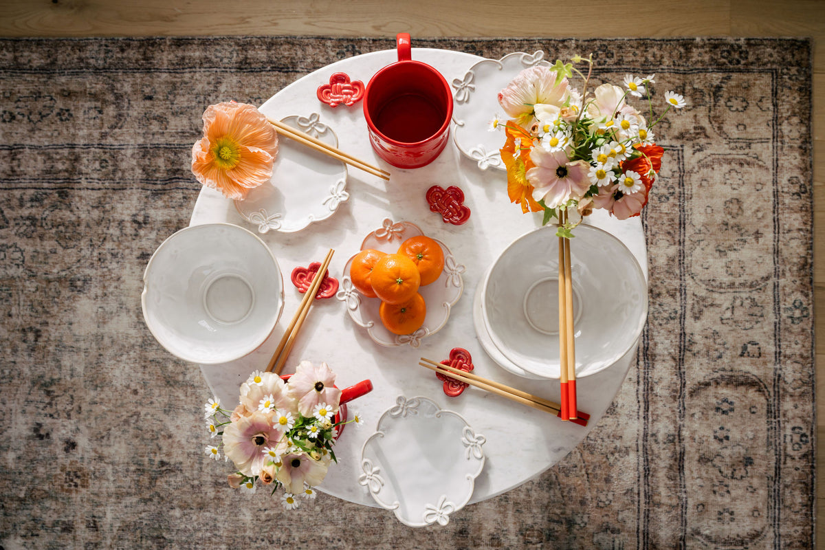 A styled overhead tabletop arrangement featuring white ceramic bowls, chopsticks with red and gold handles, a red mug, small flower bouquets, and oranges on a round white tray over a patterned rug.