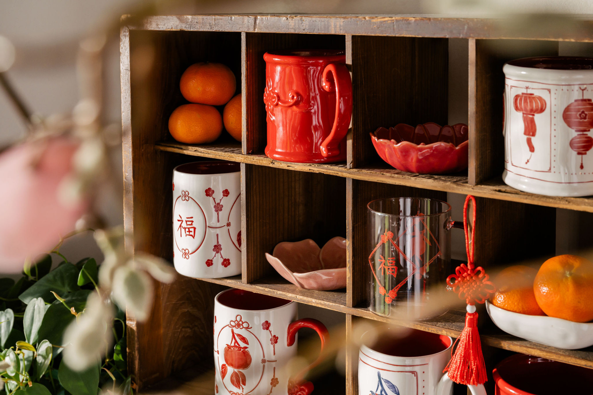 A wooden shelf filled with red and white decorative ceramic mugs, bowls, and canisters, styled with oranges and soft floral bokeh in the foreground.