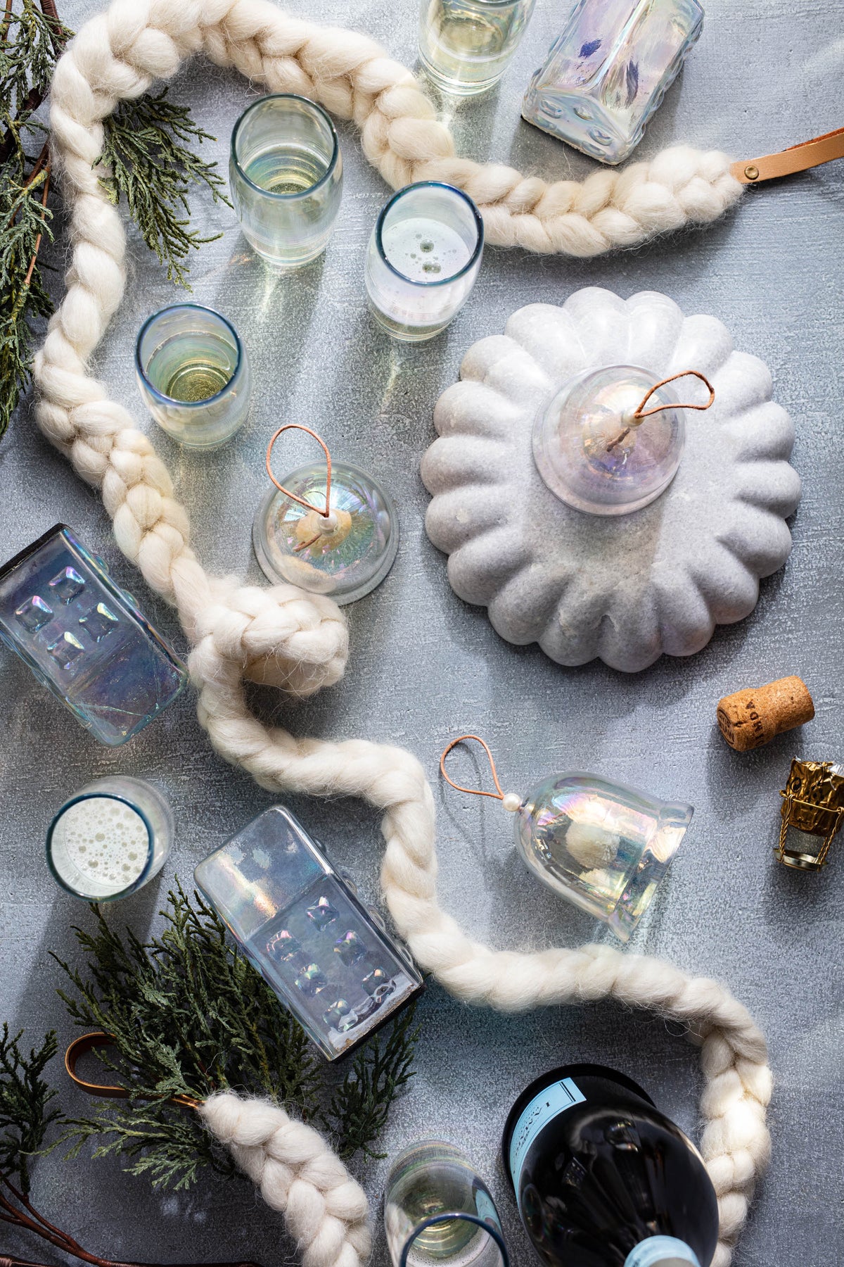 A styled flat-lay of a chunky cream knit garland draped across a gray surface with several clear glass candle holders, a white scalloped candle holder, evergreen branches, and small decorative accents.