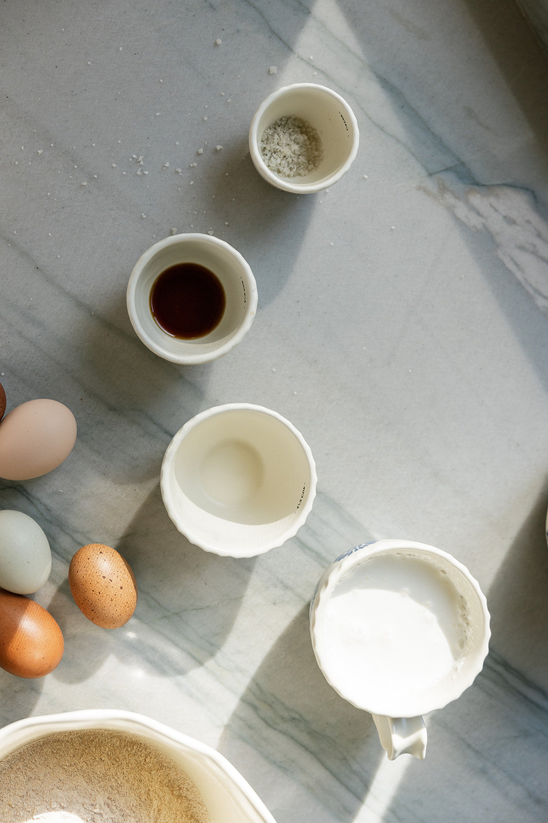 An overhead flat-lay of white ceramic cups and bowls containing ingredients, including eggs, on a light gray marble surface with natural sunlight casting shadows.