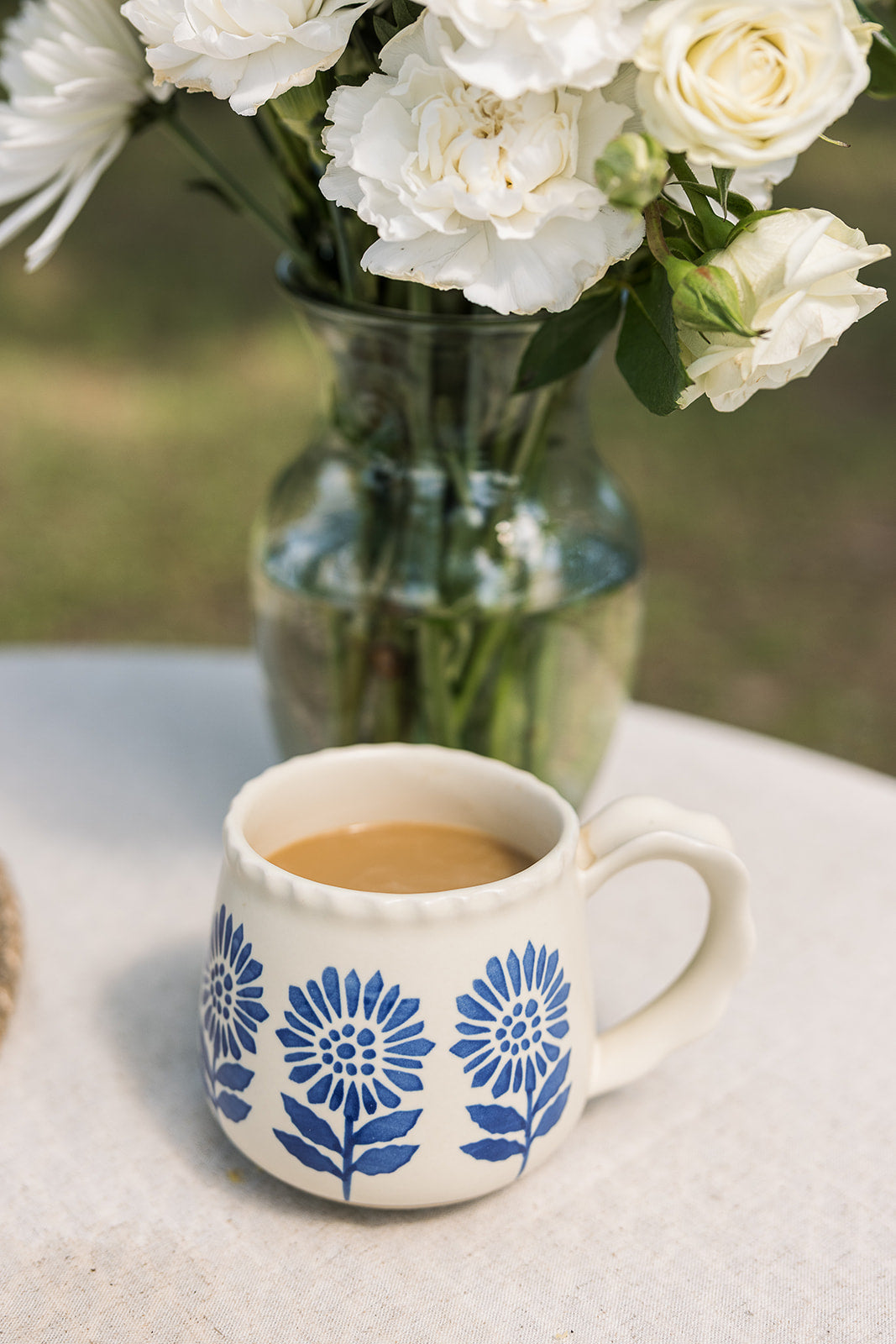 A white ceramic mug with a blue floral pattern sits on a light tablecloth, filled with a light brown beverage. In the background, a glass vase holds white flowers against a softly blurred outdoor setting.