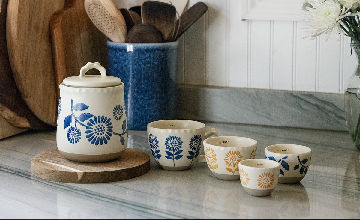 A set of ceramic kitchenware including a lidded storage jar and four small cups with floral patterns, arranged on a marble countertop in a kitchen setting with wooden cutting boards and a vase of white flowers.