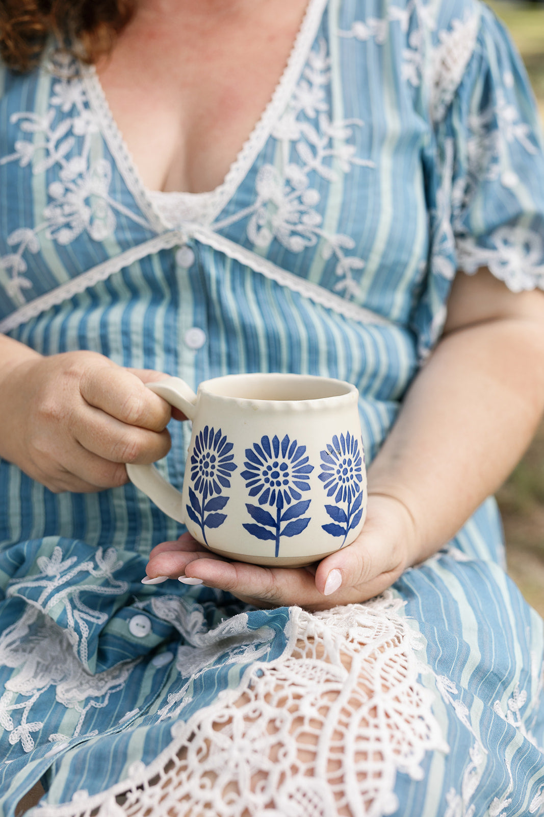 A person in a blue and white striped dress holds a cream-colored ceramic mug decorated with blue floral patterns.