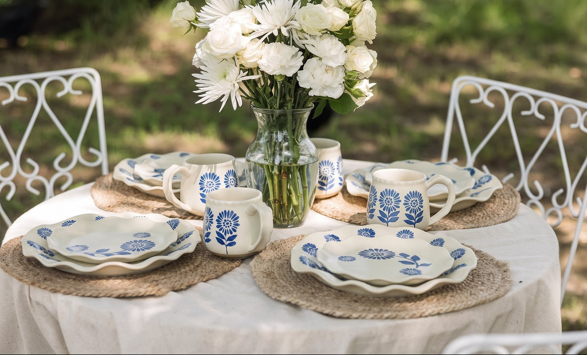 A styled outdoor dining table set with a white tablecloth, blue-and-white patterned plates, mugs, and a small cream pitcher, arranged with a glass vase of white flowers on woven placemats. White metal chairs are visible in a blurred garden background.