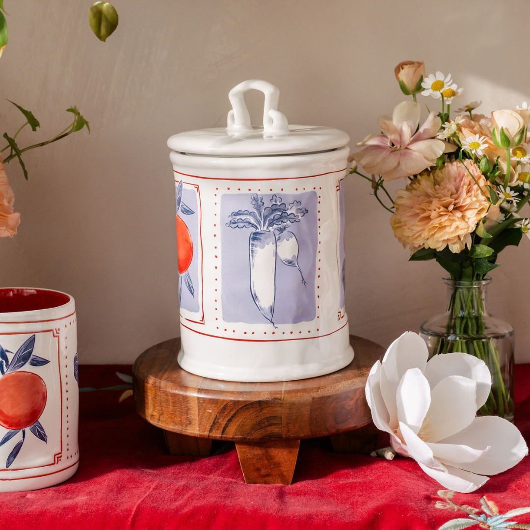 A white ceramic canister with a lid and handle, decorated with blue, red, and orange botanical illustrations, displayed on a wooden stand on a red fabric surface with flowers in the background.