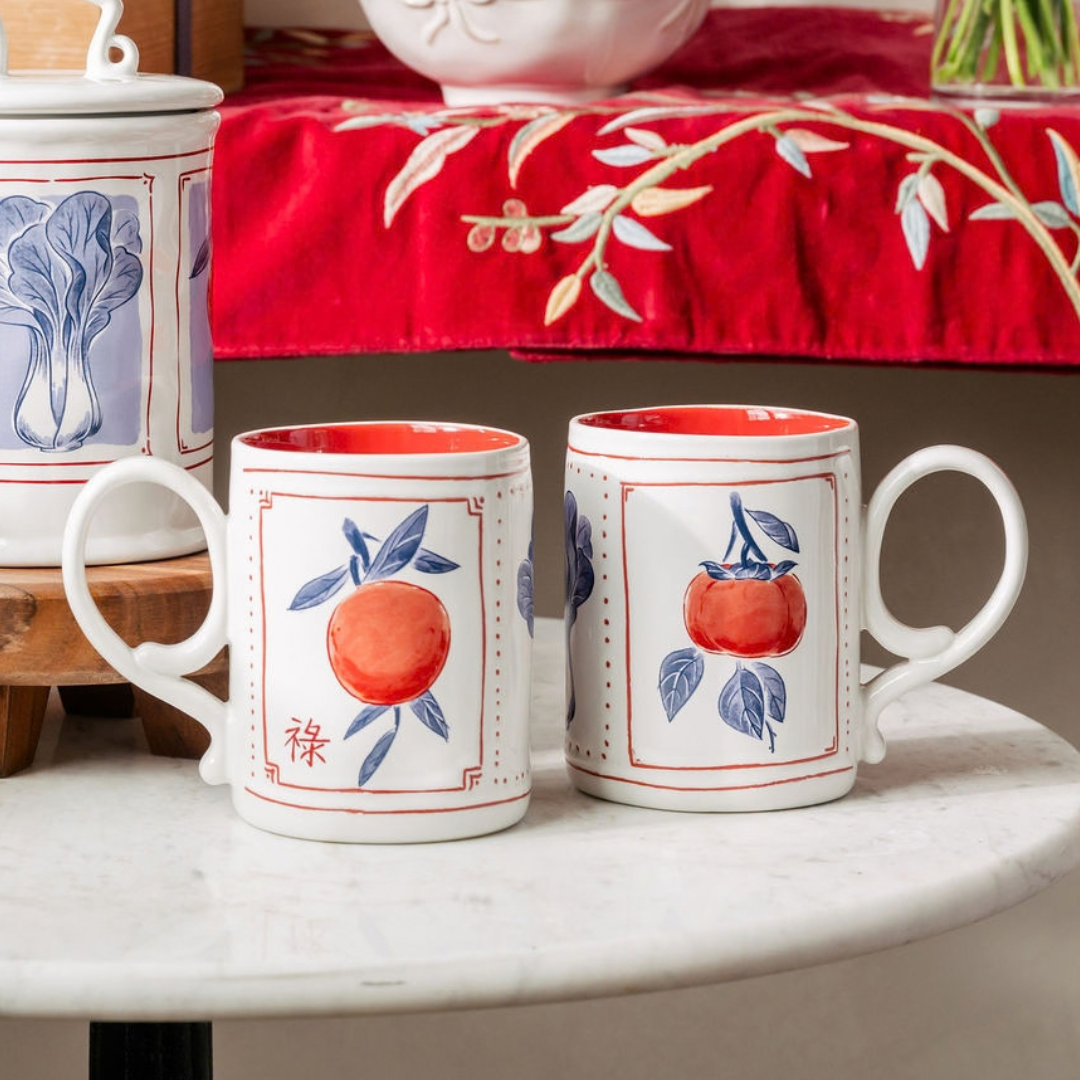 Two white ceramic mugs with red and blue persimmon illustrations, displayed on a white marble table with a red embroidered tablecloth in the background.