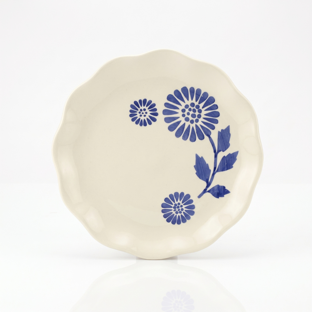 An off-white ceramic plate with a scalloped rim, decorated with blue floral patterns, shown on a clean white background with a soft reflection below.