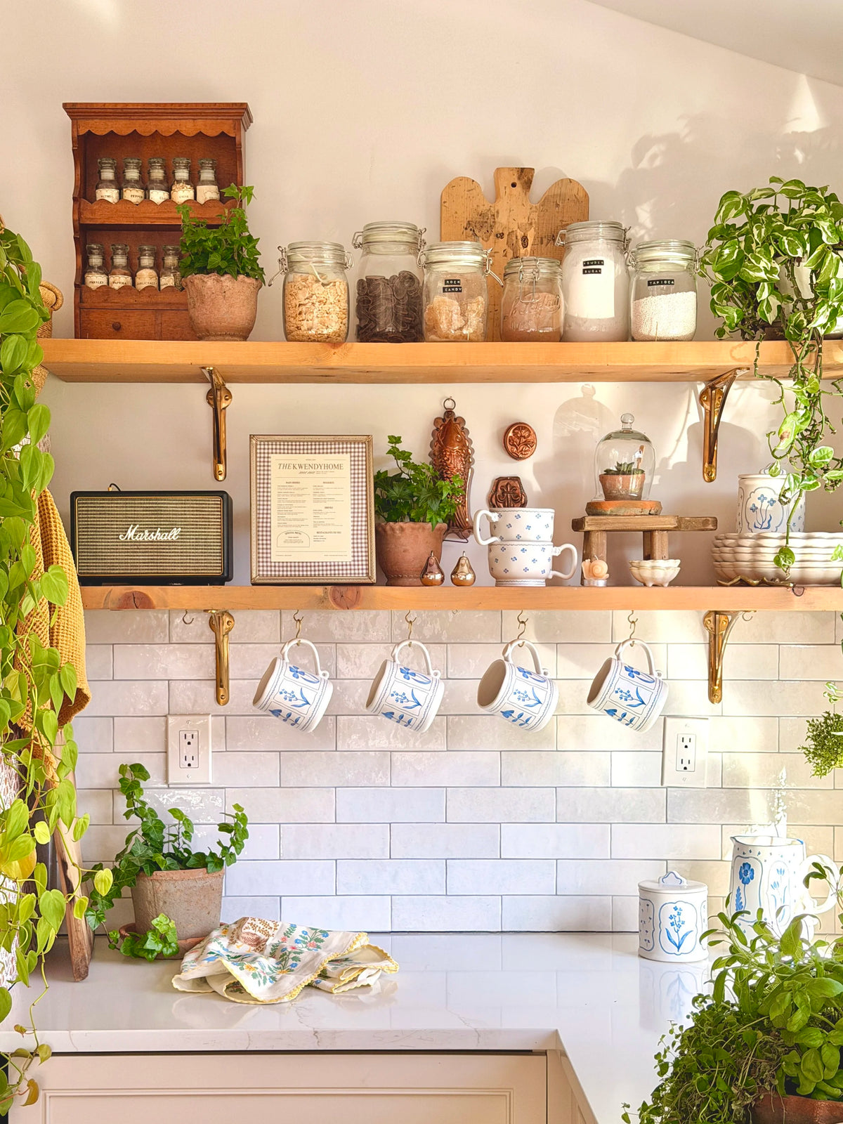 A styled kitchen shelf display featuring light wood floating shelves with brass brackets, filled with glass jars of spices and ingredients, ceramic mugs, small plants, a framed print, a vintage-style Marshall radio, and hanging white mugs against a white subway tile backsplash.