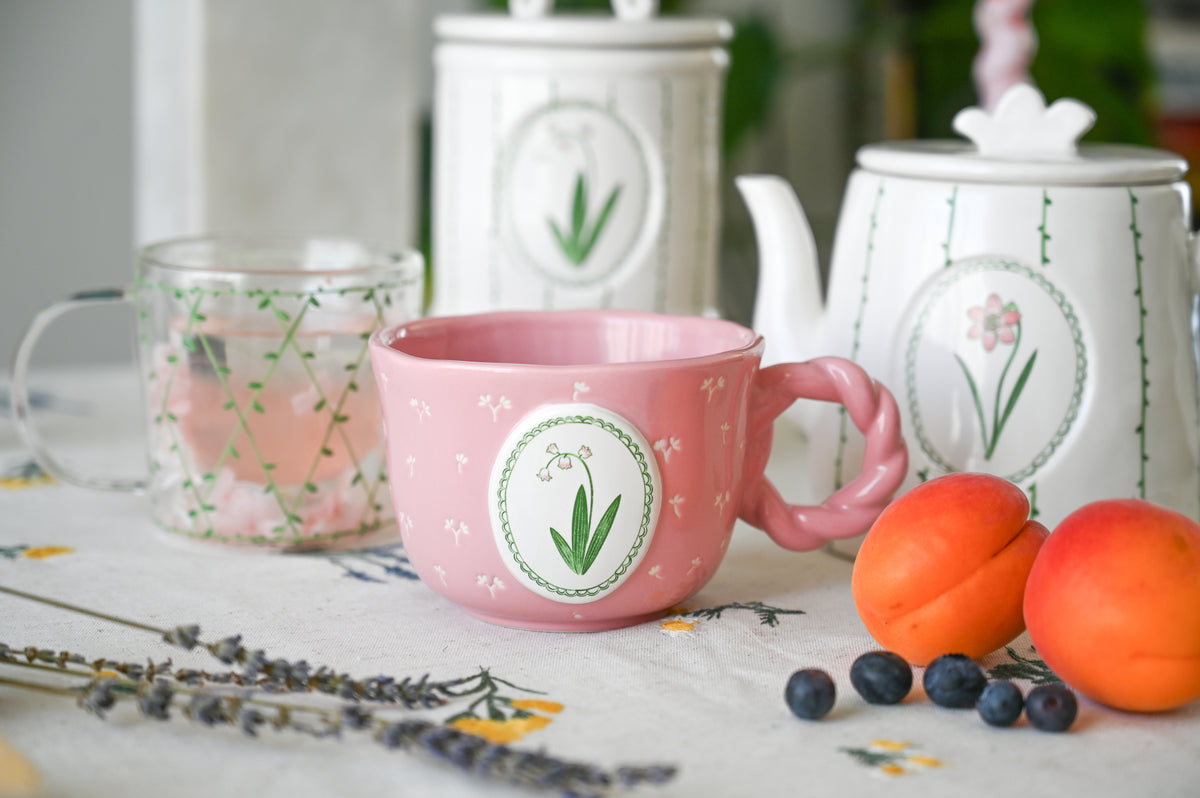 A pink ceramic mug with a floral label design sits on a tablecloth next to peaches, blueberries, and lavender, with matching white floral canisters and a teapot in the background.