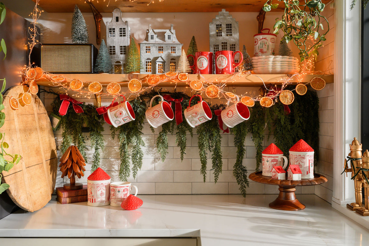 A cozy holiday kitchen shelf display featuring red and white ceramic mugs, canisters, miniature white houses, dried orange slices, green garlands, and warm string lights.