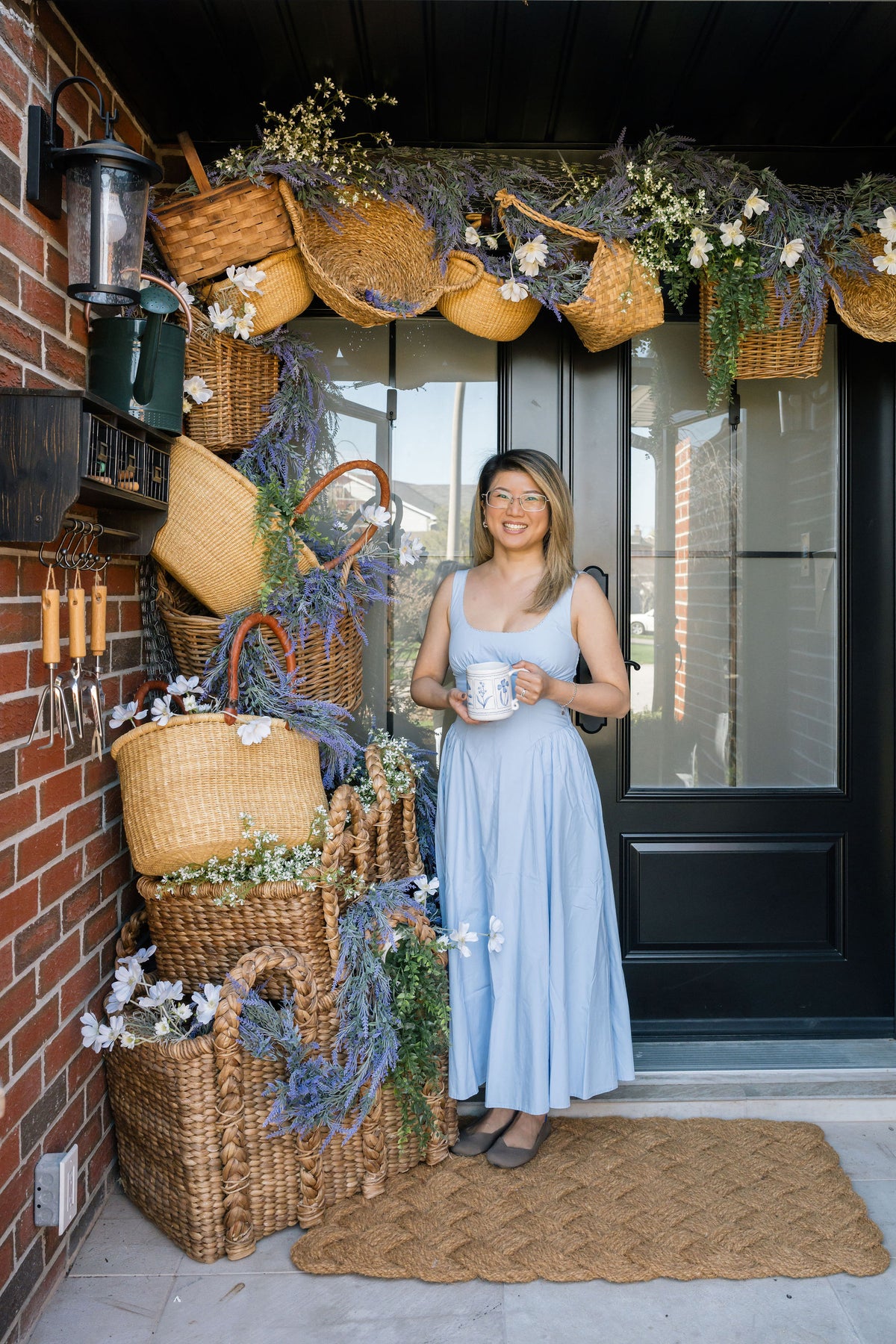 A woman in a light blue sleeveless maxi dress stands on a woven brown doormat in front of a black front door, holding a white mug. The porch is decorated with numerous woven wicker baskets, lavender sprigs, and white flowers, next to a red brick wall.