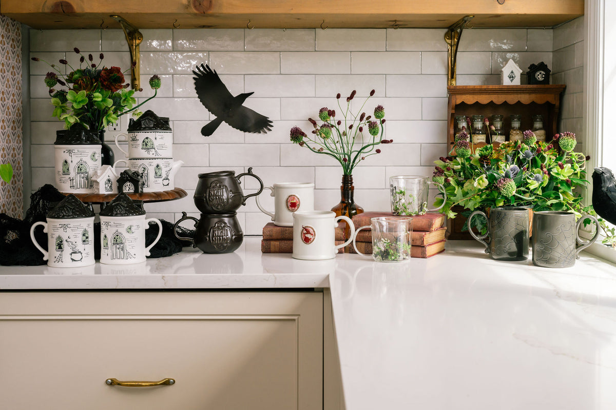 A styled kitchen countertop featuring a collection of ceramic mugs, teapots, and potted plants against a white subway tile backsplash with a wooden ceiling.