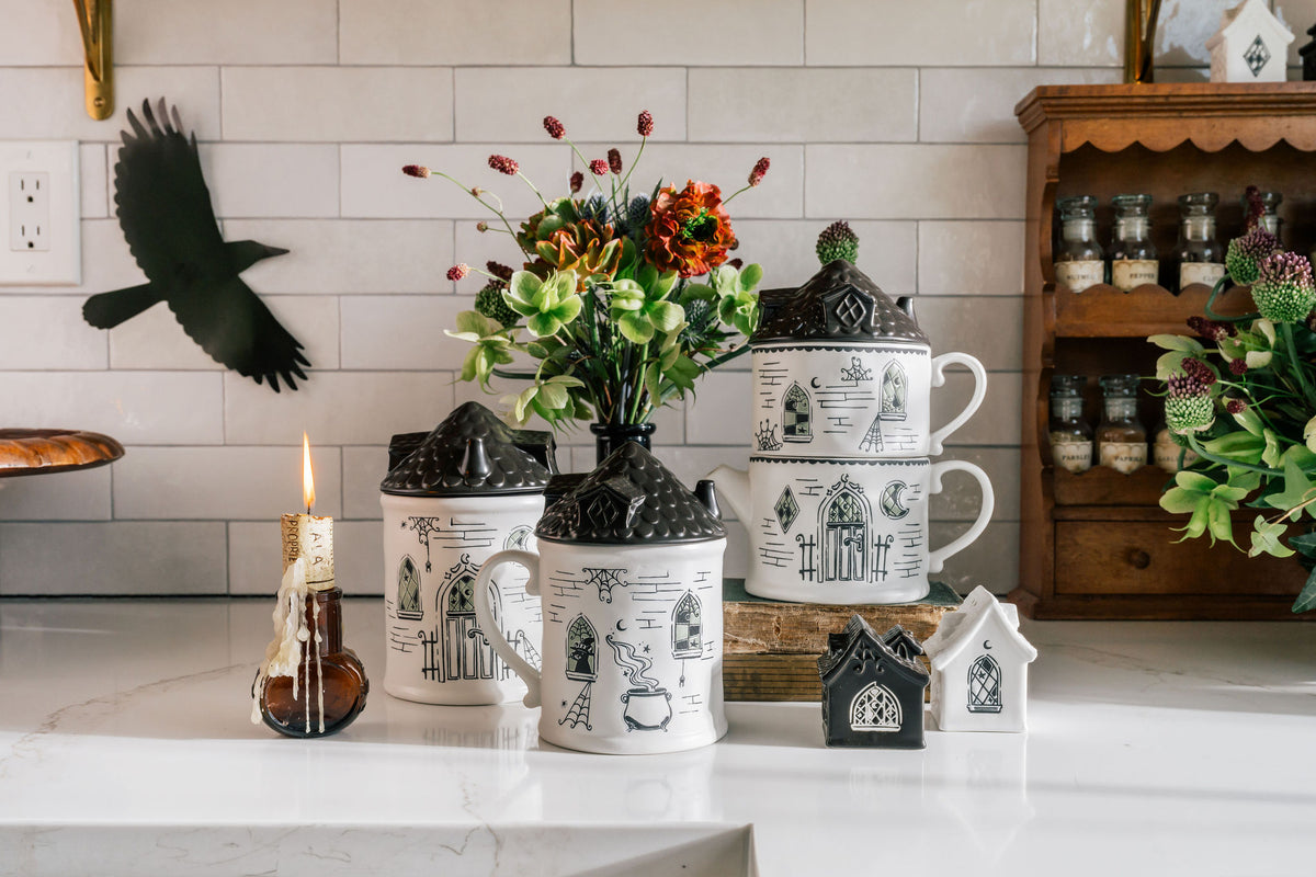 A set of black and white illustrated ceramic canisters and mugs styled on a white marble countertop in a kitchen setting, with a lit candle, flowers, and a wooden spice rack in the background.