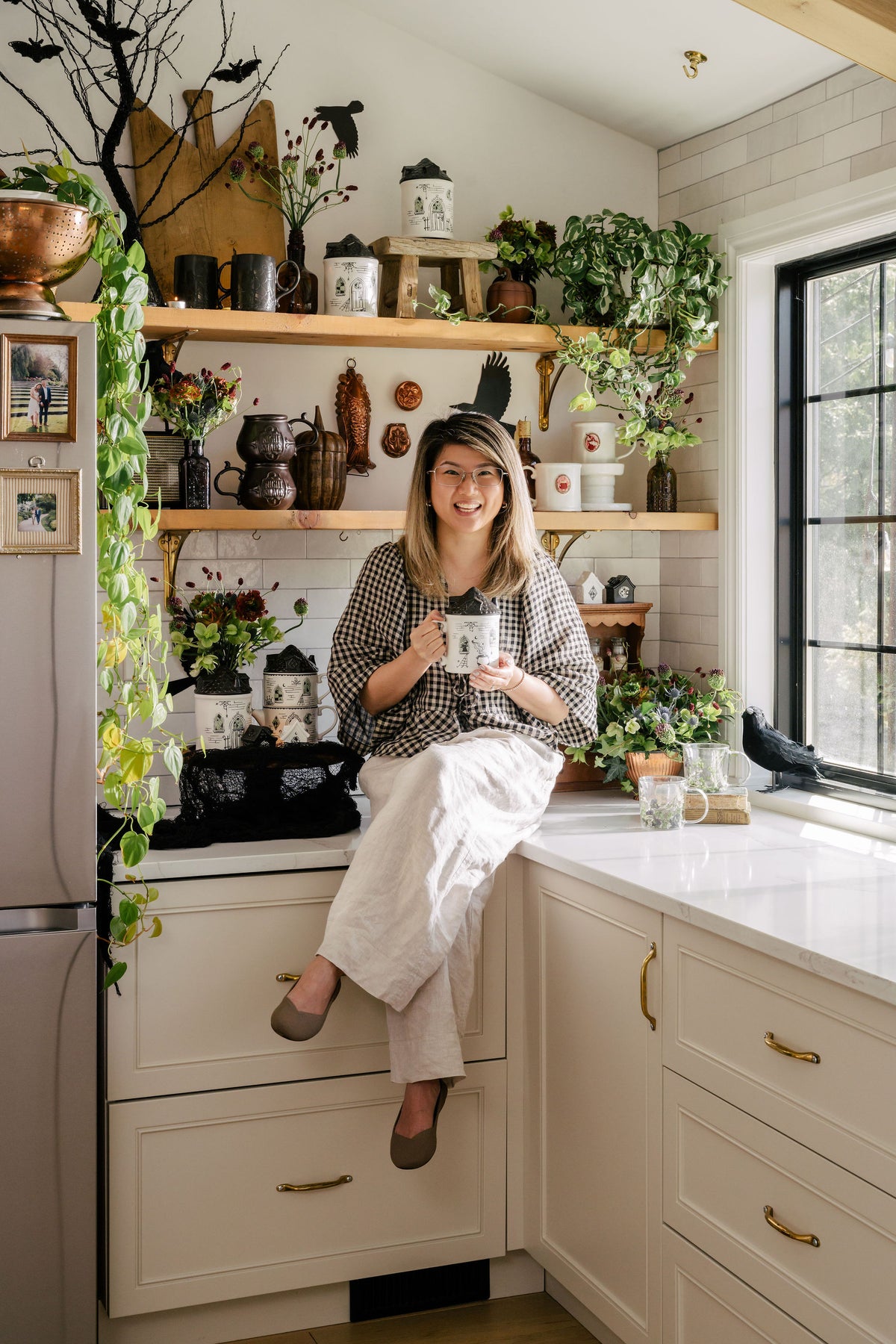 A woman wearing a black-and-white checkered top and cream pants sits on a cream kitchen cabinet, holding a white mug with a black cat perched on it. The background features a cozy, rustic kitchen with open wooden shelves, potted green plants, and brass handles.