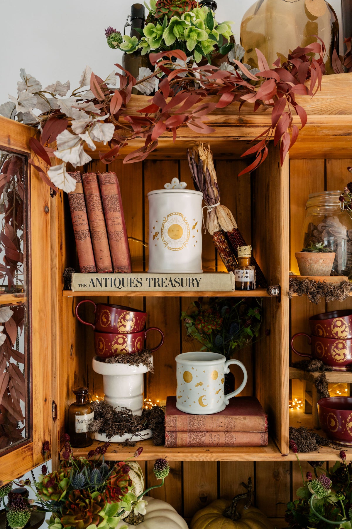 A rustic wooden display cabinet styled with autumn decor, featuring vintage-style ceramic mugs, books, a white canister labeled 'ANTiques TREASURY,' dried leaves, small bottles, and warm string lights.