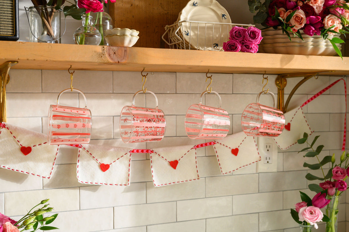 A kitchen shelf decorated with four clear plastic cups filled with pink liquid, hanging from gold hooks above a white subway tile backsplash. A white fabric banner with red hearts and red dashed borders hangs below.