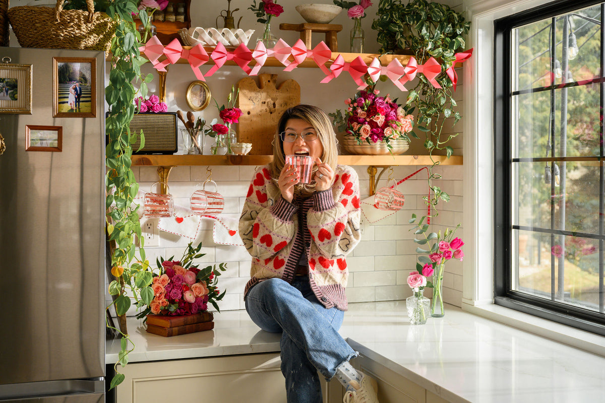 A woman wearing a red and white heart-patterned knit cardigan sits on a kitchen counter, holding a mug. The kitchen is decorated with pink paper garlands, hanging stockings, and various flowers.