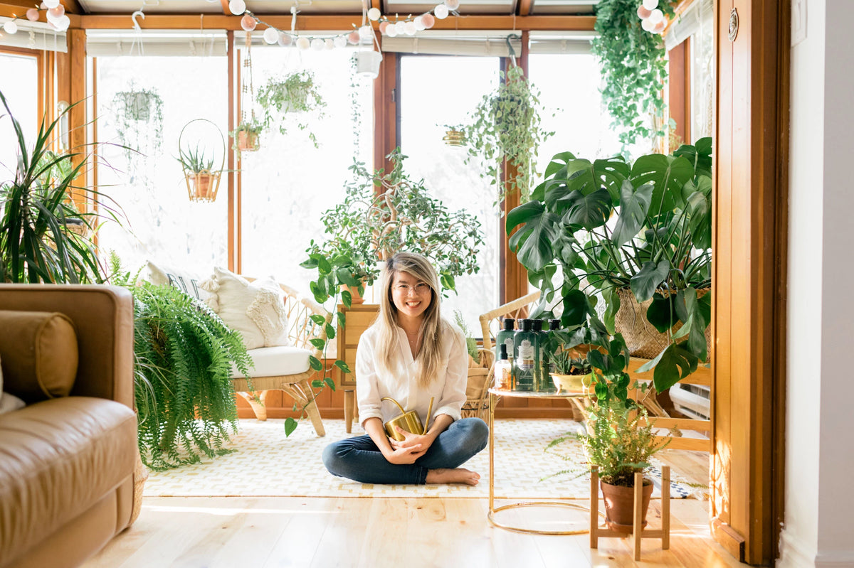 A woman sits cross-legged on a light rug in a bright, plant-filled room with large windows, wooden trim, and indoor plants. She is wearing a light beige top and dark blue jeans, holding a small potted plant.