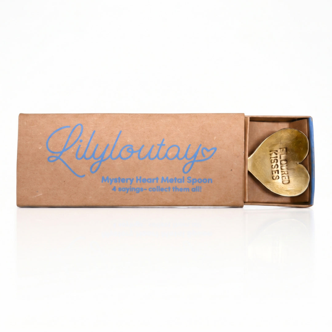 A brown cardboard matchbox-style package with blue script reading 'Lilyloutray' and smaller text 'Mystery Heart Metal Spoon' and '4 spoons - collect them all!' sits open on a white background, revealing a gold heart-shaped spoon inside with 'KISSES LILYLOUTAY' printed on it.
