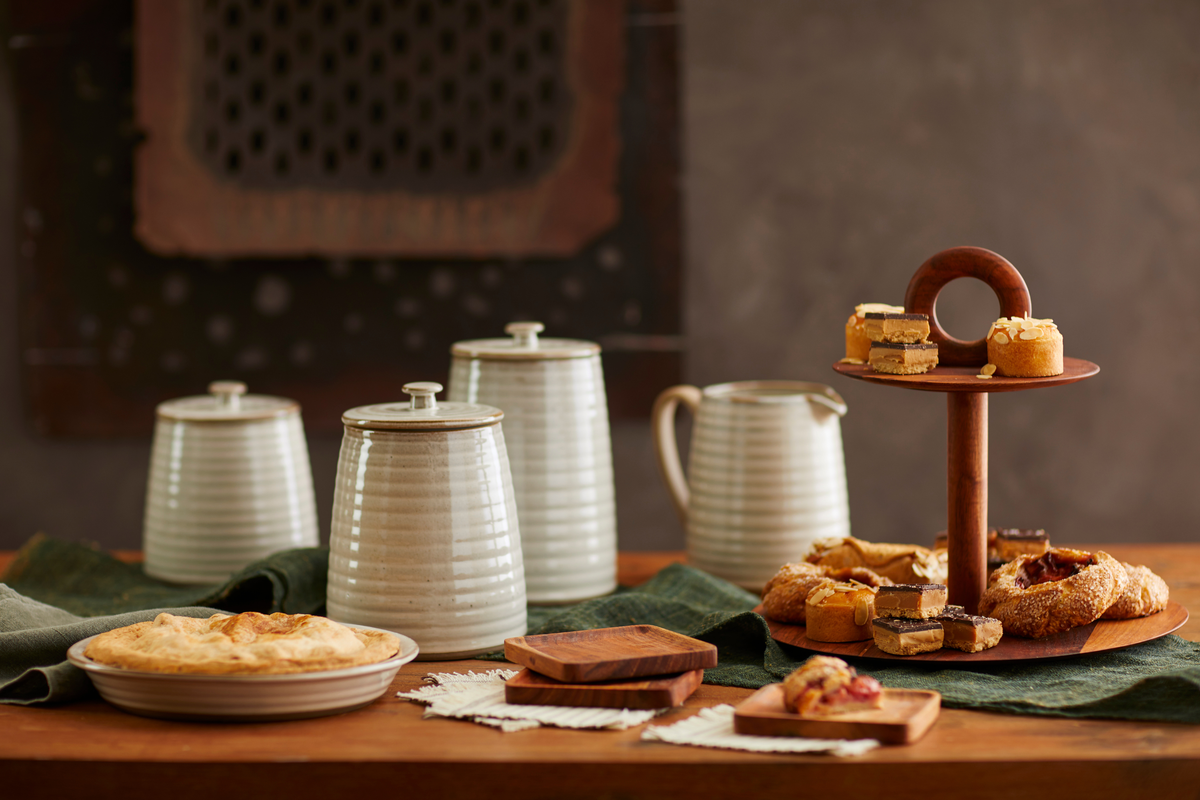 A styled tea or dessert spread on a wooden table with cream-colored ribbed ceramic canisters, a small pitcher, a two-tier wooden stand holding pastries, and a pie in the foreground against a warm brown background.