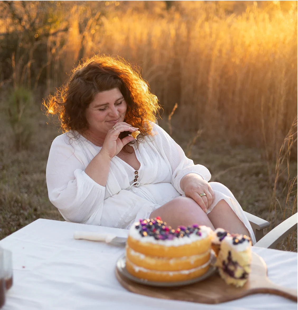 A woman in a white dress sits outdoors at sunset beside a table with a sliced cake and a donut, with tall dry grass in the background.