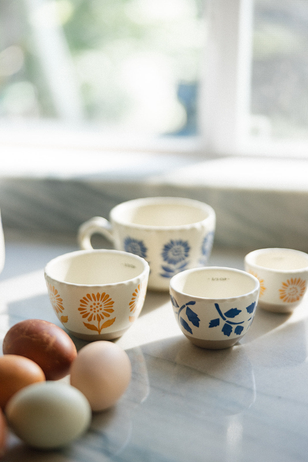 A set of ceramic cups with floral patterns in orange and blue, arranged on a marble countertop with eggs in the foreground and a softly blurred kitchen background.