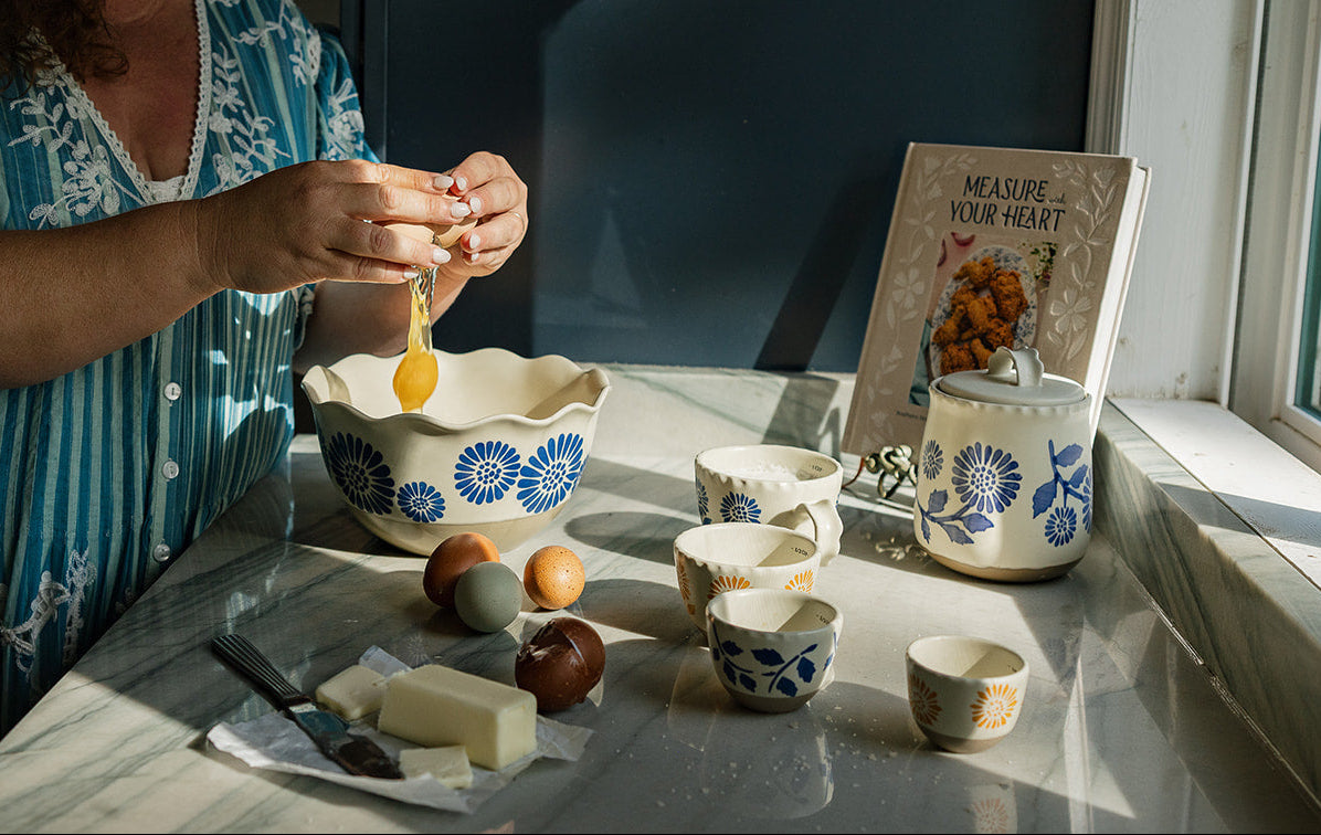 A woman in a blue and white patterned dress cracks an egg into a matching blue and white ceramic bowl on a marble countertop. Several small ceramic dishes, a cookbook titled 'Measure Your Heart,' and food items are arranged nearby in a sunlit kitchen setting.