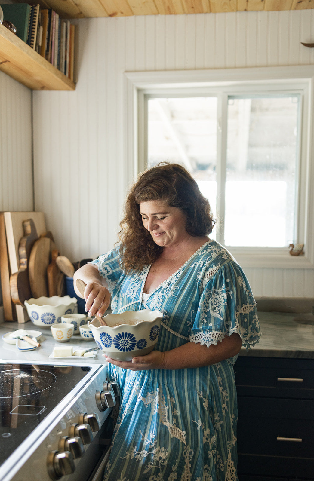 A woman with curly brown hair wearing a blue and white patterned dress stands in a kitchen, holding a white bowl with blue floral patterns while mixing a bowl with a wooden spoon.