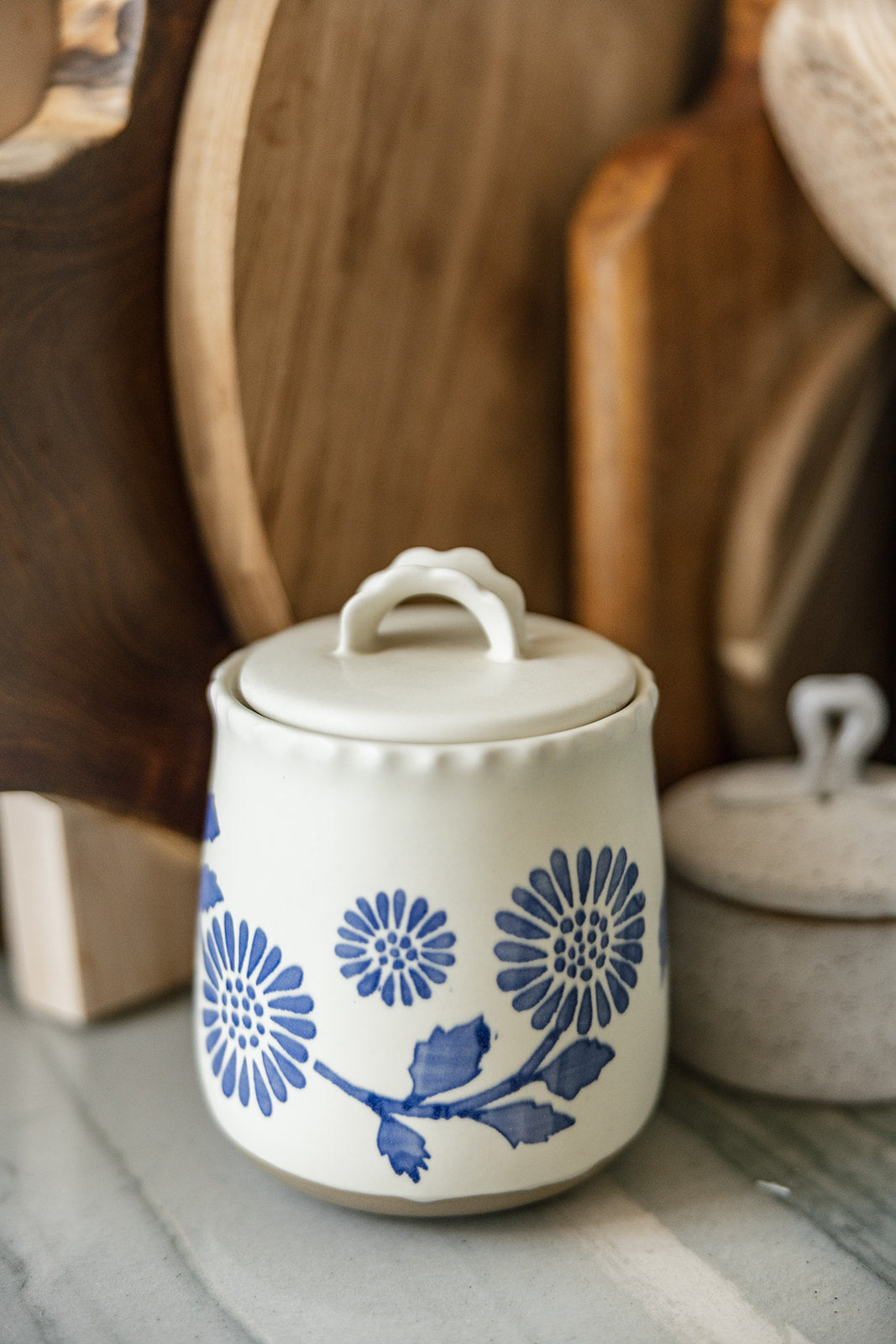 A small white ceramic lidded jar with a blue floral pattern, set on a marble surface with rustic wooden cutting boards in the background.