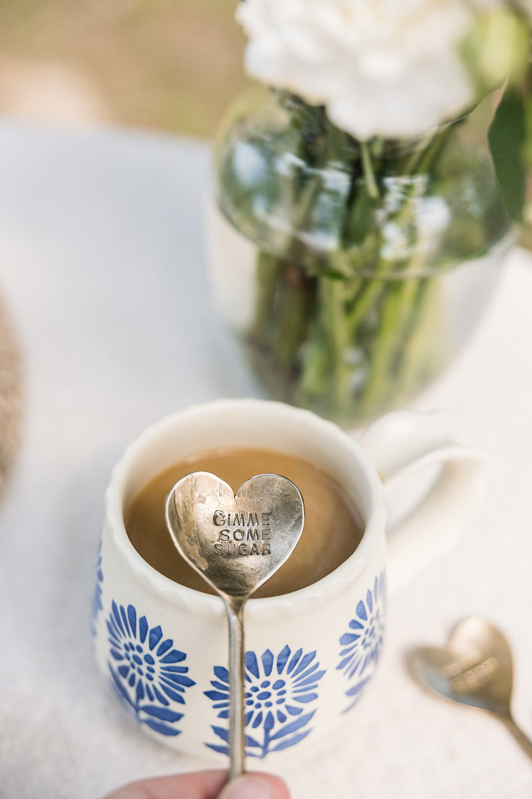 A white ceramic mug with blue floral patterns filled with coffee, holding a heart-shaped metal spoon engraved with 'GIVE SOME BACK'. A hand is holding the spoon, with a blurred glass vase of white flowers in the background.