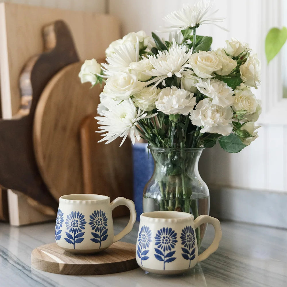 Two cream ceramic mugs with blue floral patterns sit on a marble countertop next to a glass vase filled with white flowers, with wooden cutting boards in the background.