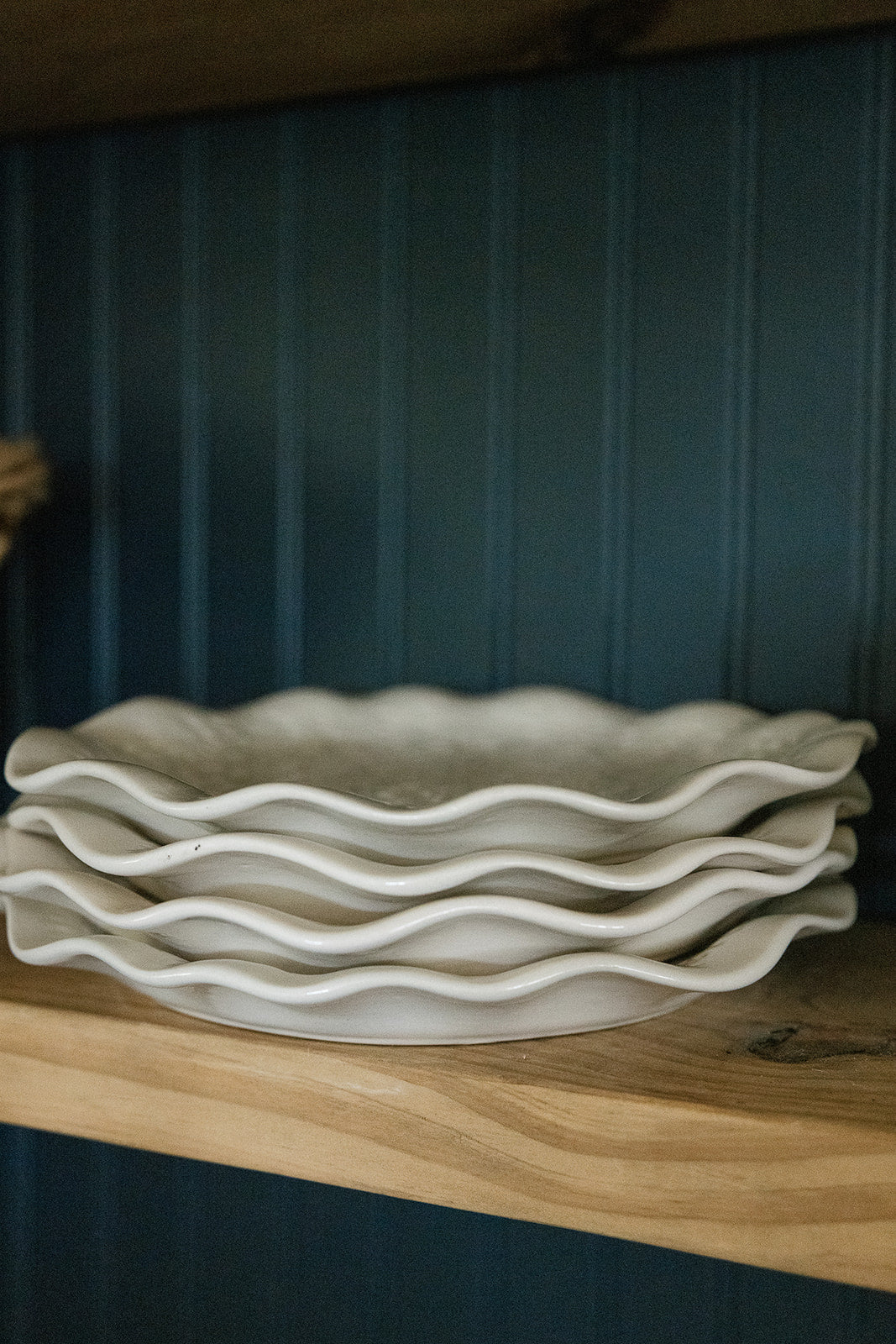 A stack of four white ceramic wavy-edged bowls displayed on a light wood shelf against a dark blue paneled background.
