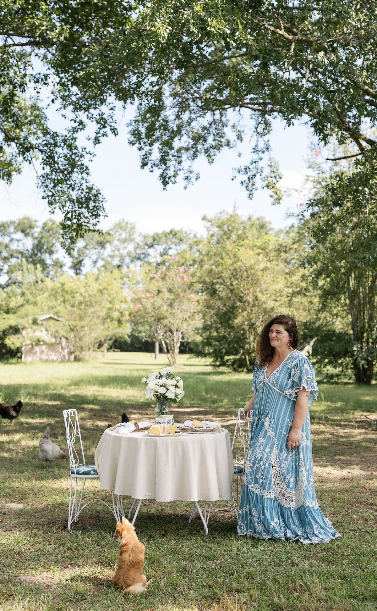 A woman wearing a light blue patterned bohemian maxi dress stands next to an outdoor dining table set on a grassy lawn under a large tree, with a ginger cat sitting in the foreground.