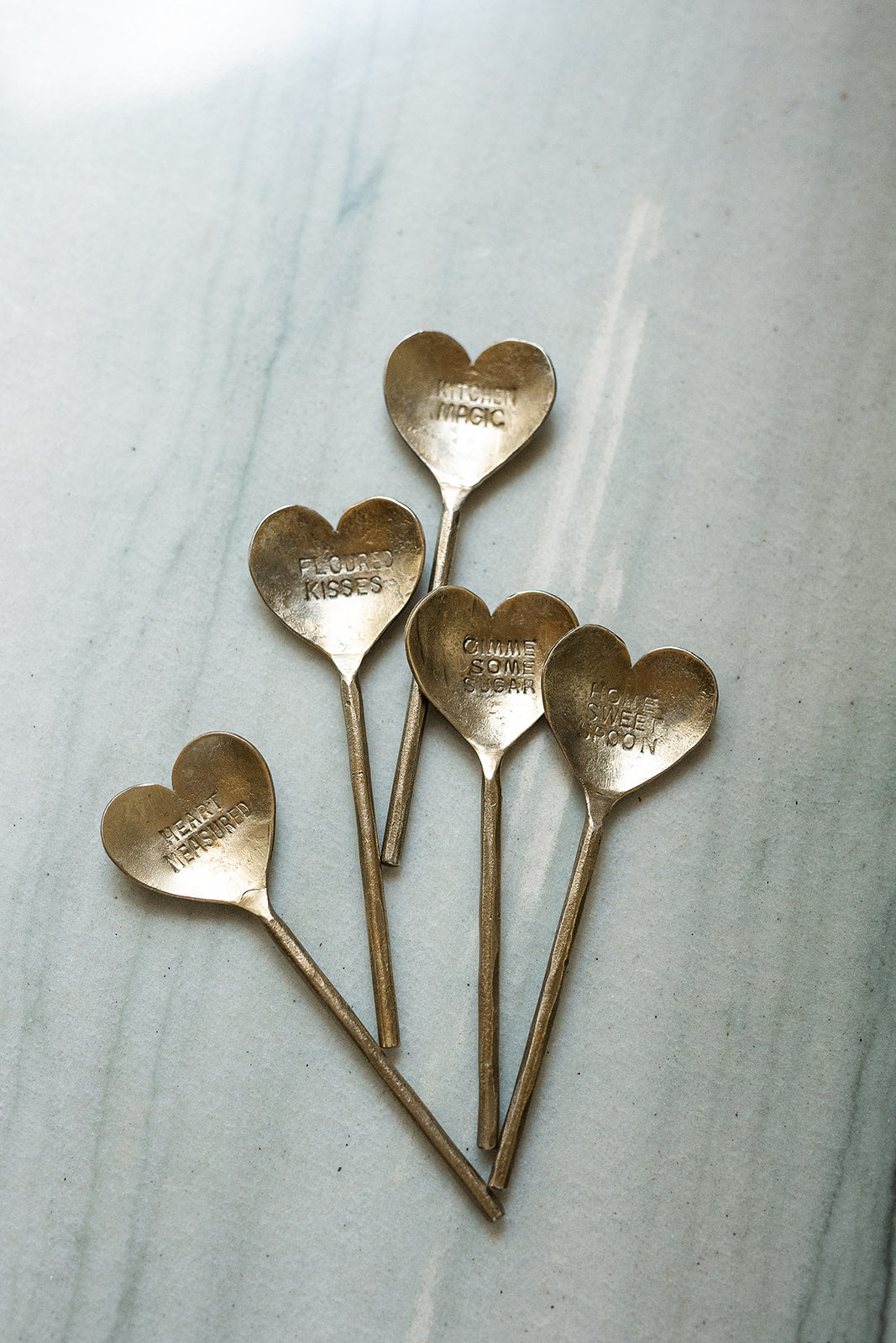 Five small heart-shaped metal spoons with engraved names and 'LOVE YOU' on the bowls, arranged on a light marble surface.