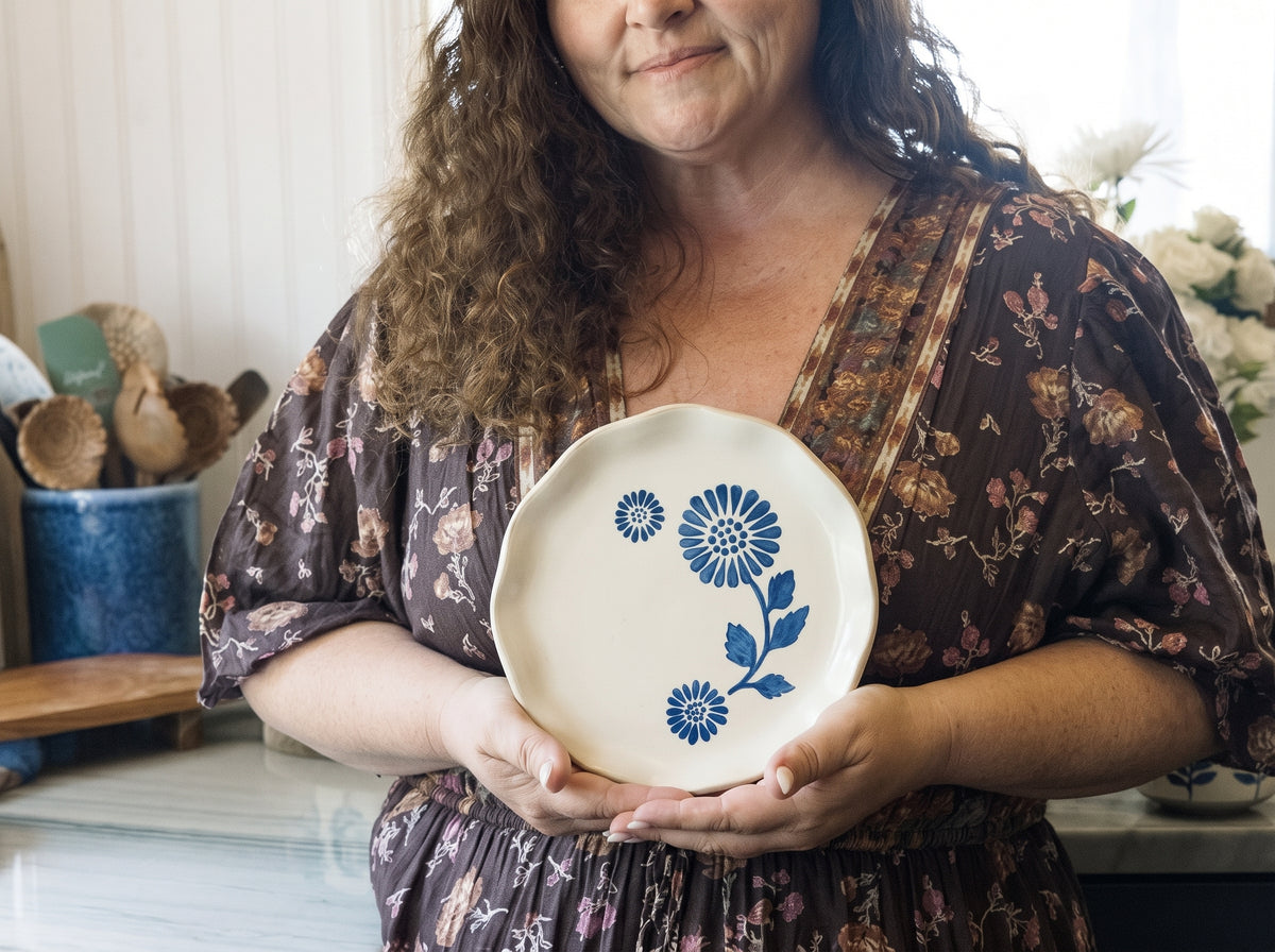 A woman in a brown floral dress holds a round off-white ceramic plate decorated with blue floral patterns, set against a softly blurred kitchen background.